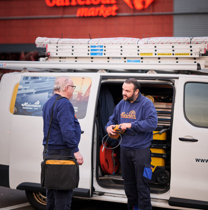 Zwei Handwerker stehen vor einem weißen Transporter mit Leitern auf dem Dach vor einem Carrefour Supermarkt in Belgien. Sie besprechen Elektroinstallationen im Supermarkt, wobei Werkzeuge und Ausrüstung für Schaltanlagen und Niederspannungsanlage sichtbar sind. Die Szene symbolisiert Not- und Sicherheitsbeleuchtung im Einzelhandel, Metallelemente im Geschäft sowie Einbruchschutz für Supermärkte und Kommunikationsnetze im Einzelhandel.