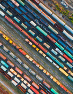 Luftaufnahme eines Industrie-Güterbahnhofs mit zahlreichen farbigen Eisenbahnwaggons auf mehreren Gleisen, umgeben von Infrastruktur und Grünflächen, ideal für Planung und Entwicklung sowie elektrotechnische Beratung und Umsetzung in Industrie, öffentliche Gebäude und Infrastrukturprojekten.