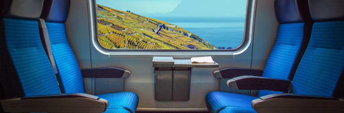Blick aus einem Zugabteil mit blauen Sitzen auf eine malerische Landschaft mit Weinbergen, See und Bergen. Dieses Bild symbolisiert die Planung und Entwicklung moderner Infrastruktur im Bereich öffentlicher Gebäude und Industrie sowie die elektrotechnische Beratung und Umsetzung in Verkehrsmitteln.