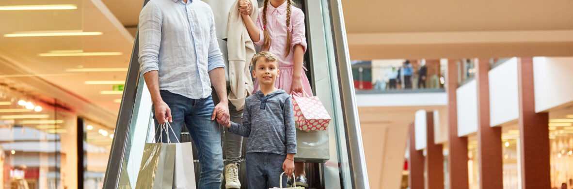 Eine Familie mit zwei Kindern fährt in einem modernen Einkaufszentrum auf einer Rolltreppe nach unten. Sie trägt Einkaufstüten und befindet sich in einem Geschäftsumfeld, das typische Sektoren wie Einzelhandel und Großhandel bedient. Die Infrastruktur des Gebäudes ist hell und offen gestaltet, was auch für öffentliche Gebäude und öffentliche Bauprojekte charakteristisch ist. Die Elektrotechnik und Produktionsanlagen im Hintergrund sind nicht sichtbar, jedoch impliziert die moderne Architektur eine hochwertige technische Ausstattung.
