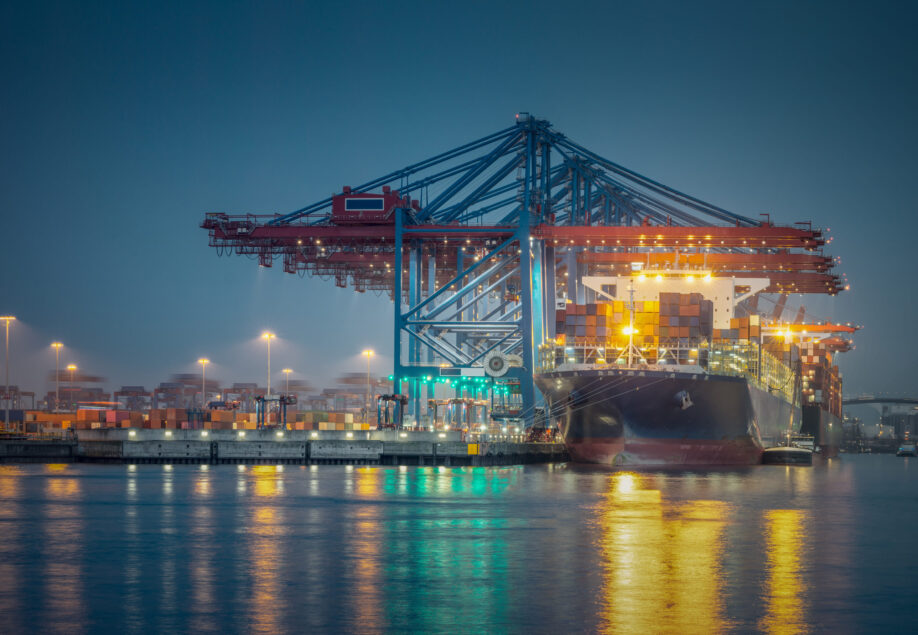 Hafen mit großen Containerschiffen und Produktionsanlagen bei Nacht, beleuchtet und mit Kränen für den Umschlag von Gütern. Die Szene zeigt wichtige Infrastruktur für den Großhandel und Einzelhandel sowie öffentliche Bauprojekte. Die Elektrotechnik der Sektoren sorgt für reibungslose Abläufe in diesem Industrie- und Geschäftsumfeld nahe öffentlicher Gebäude.