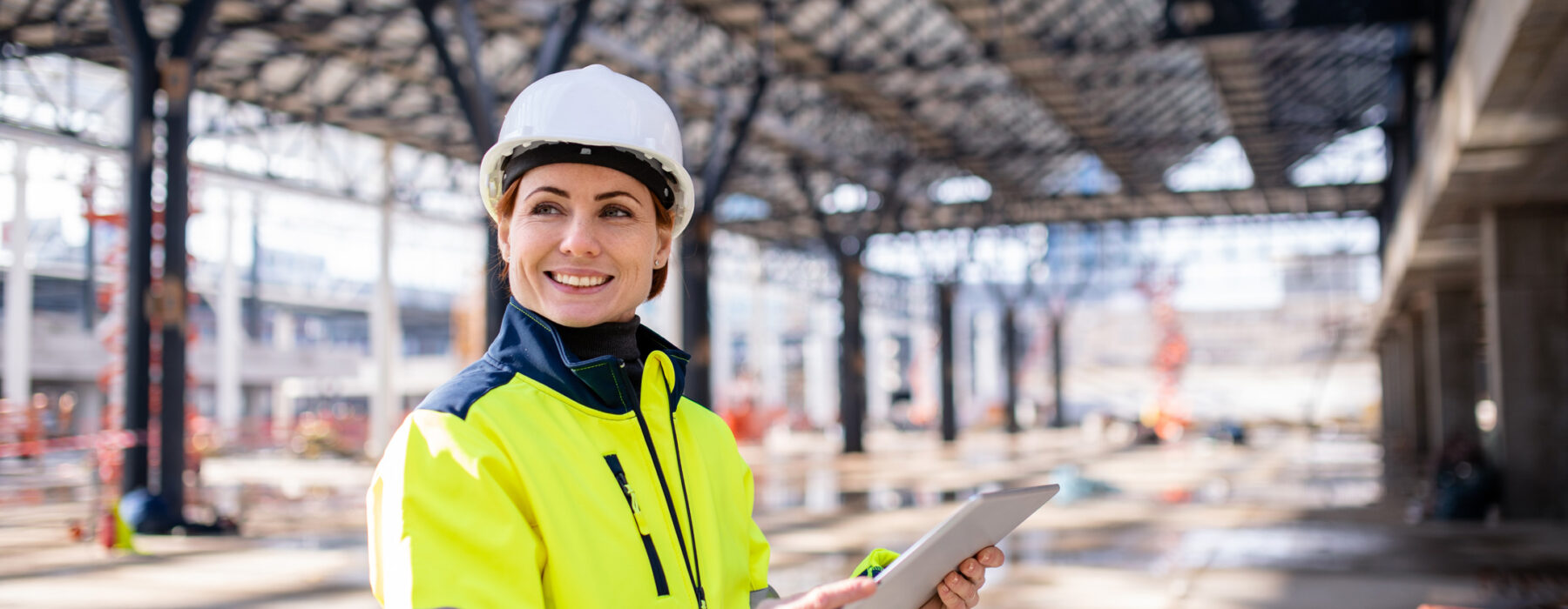 Eine Frau in Schutzhelm und leuchtend gelber Arbeitsjacke steht auf einer Baustelle und hält ein Tablet. Sie ist in der Planung und Entwicklung sowie der elektrotechnischen Beratung tätig und überwacht die Umsetzung von Projekten in der Industrie, öffentlichen Gebäuden und Infrastruktur.