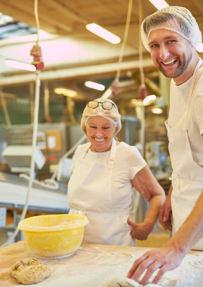 In einer modernen Produktionsanlage der Lebensmittelindustrie arbeiten zwei Bäcker in Schutzkleidung an einem Holztisch mit Teig. Die Anlage ist mit fortschrittlicher Elektrotechnik ausgestattet und gehört zu den wichtigen Sektoren der Industrie. Solche Produktionsanlagen sind essenziell für den Großhandel, Einzelhandel und das Geschäft. Öffentliche Gebäude und Bauprojekte profitieren ebenfalls von der Infrastruktur, die durch innovative industrielle Lösungen unterstützt wird.