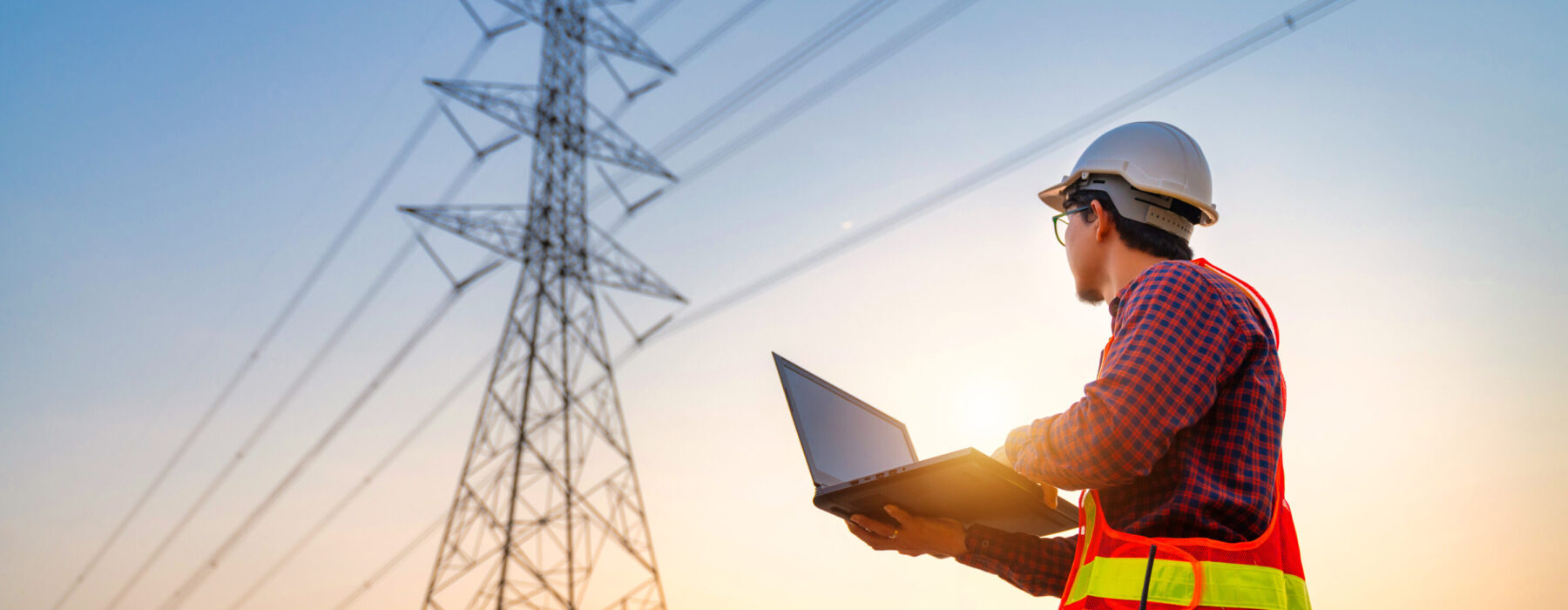 Ein Techniker im Industrie Bereich führt Elektro Arbeiten an Mittel- und Hochspannungsanlagen durch. Er trägt einen Schutzhelm und eine Warnweste und überprüft mit einem Laptop die Verkabelungssysteme an einem Hochspannungsmast. Im Hintergrund sind Stromleitungen zu sehen, die für Schalttafeln, Schaltschränke und Glasfaserarbeiten relevant sind. Die Szene symbolisiert auch den Einsatz erneuerbarer Energien in der modernen Energieversorgung.