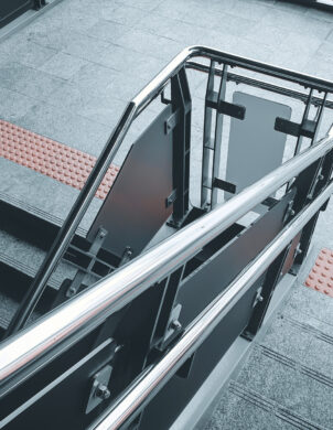 Moderne Treppe mit Edelstahlgeländer und taktilen Leitsystemen aus orangenen Noppenplatten in einem öffentlichen Gebäude, typisch für Infrastrukturprojekte und öffentliche Bauprojekte. Die robuste Bauweise und Elektrotechnik-Elemente sind geeignet für verschiedene Sektoren wie Industrie, Produktionsanlagen, Geschäft, Einzelhandel und Großhandel.