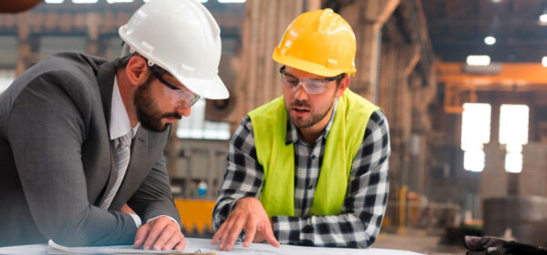 Zwei Fachleute in einem Industrieumfeld besprechen gemeinsam Baupläne. Einer trägt einen Anzug und weißen Helm, der andere eine Sicherheitsweste und gelben Helm. Die Szene symbolisiert Planung und Entwicklung sowie elektrotechnische Beratung und Umsetzung in Industrie, öffentlichen Gebäuden und Infrastrukturprojekten.
