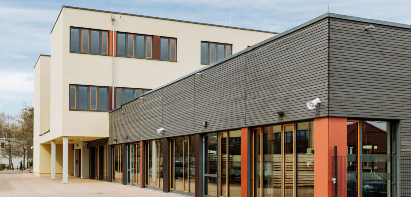 Modernes Gebäude mit klarer Architektur, bestehend aus einem mehrstöckigen hellen Baukörper und einem eingeschossigen Anbau mit dunkler Holzverkleidung und großen Fenstern. Die Fassade zeigt Elemente der Elektrotechnik mit Außenleuchten und Sicherheitskameras. Dieses Bauwerk eignet sich für verschiedene Sektoren wie Industrie, Produktionsanlagen, Geschäft, Einzelhandel und Großhandel. Es kann zudem als Teil von öffentlichen Gebäuden, öffentlichen Bauprojekten und Infrastruktur genutzt werden.