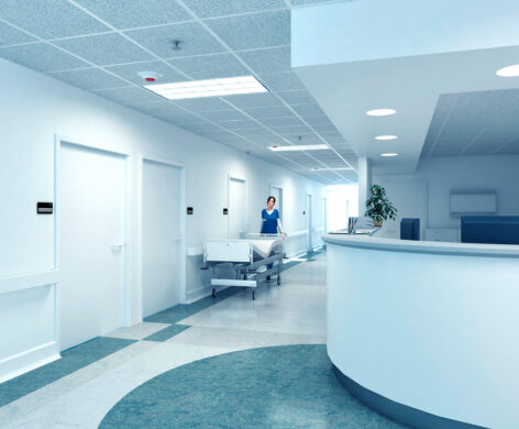 Moderner, sauberer Krankenhausflur mit mehreren weißen Türen, einer Empfangstheke und einer Pflegekraft, die ein Krankenhausbett schiebt. Die helle Beleuchtung und die strukturierte Decke zeigen hochwertige Elektrotechnik. Dieses Bild steht beispielhaft für öffentliche Gebäude und öffentliche Bauprojekte im Bereich Infrastruktur. Die Ausstattung und Organisation spiegeln Anforderungen aus verschiedenen Sektoren wider, darunter Industrie, Produktionsanlagen sowie Geschäft, Einzelhandel und Großhandel.